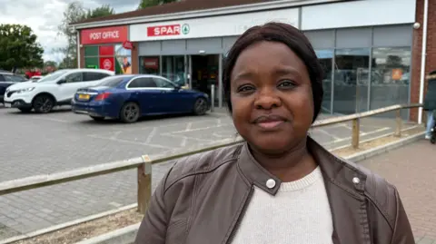 Ben Schofield/BBC Avese Dio, standing outside shops, with two cars outside, the woman is wearing a brown jacket, beige top, 