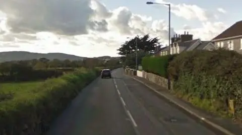The main road through Colby. There are fields on the left and houses on the right, with a car driving away on the road in the distance