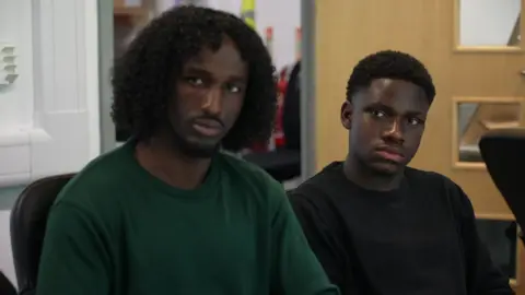 Adnan, 18, wears a dark green top and is sitting at a table inside an office room beside Edem, 18, in a black top, both listening to the discussion 