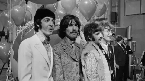 Getty Images The Beatles are pictured lined up together during press call at Abbey Road Studios in a black and white photo. There are balloons behind them and music recording equipment. They are wearing jackets and multi-patterned shirts.