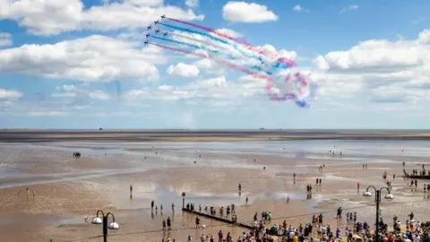 MOD Hundreds of people watch the red arrows above the beach at Cleethorpes. The tide is out so far you can hardly see the sea, and the sand is wet with pooling water.