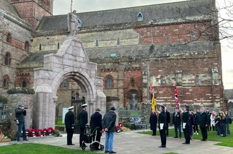 Two flagbearers are are joined by veterans and members of the public outside St Magnus Cathedral. THey are standing in from of a stone arched gateway marked with the dates of WW1 and inscribed with the names of those who did not return from the war.