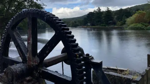 The picture looks out over the River Derwent in Derbyshire. The water is calm and there are clouds and some blue in the sky. In the foreground there is a large, old cog wheel from the days when Belper Mill was in use. 