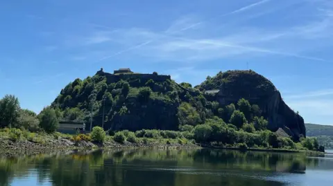 Brian Watt Two steep hills of Dumbarton Rock covered in bushes and trees in the sunshine. The sky is blue and vapour trails 