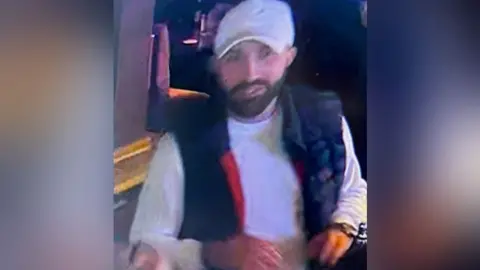 Avon and Somerset Police CCTV footage of a man wearing a white jumper, black vest and white baseball cap. He has a dark beard and is inside a venue.