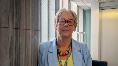A woman with short blonde hair is wearing a blue blazer and a yellow blouse. She is looking towards the camera with a slight smile.