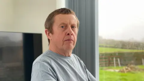 A fair-haired man wearing a blue t-shirt sat in front of a large window through which fields and a wooden fence can be seen.