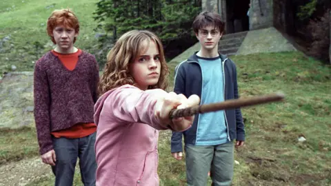 Murray Close/Getty Images On the set of the film Harry Potter and the Prisoner of Azkaban, Hermione looks serious while pointing her wand. Ron and Harry stand on the grassy hill behind her.