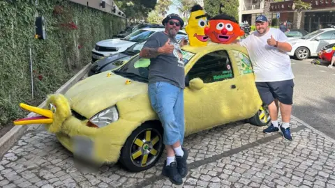 A furry yellow car decorated to look like Big Bird from Sesame Street is parked in a European street. Two men wearing shorts and T-shirts are standing next to it, giving the thumbs-up and smiling. 