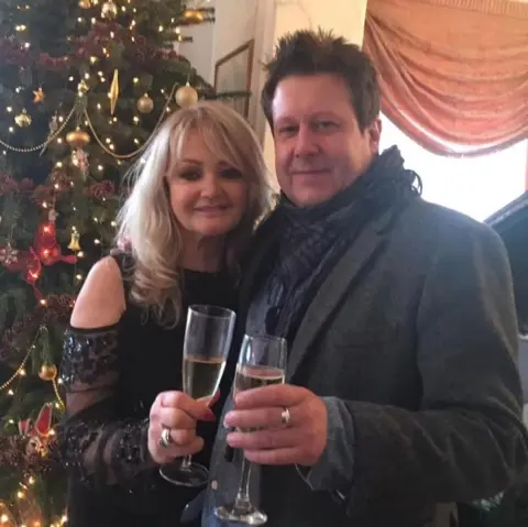 Rob Marshall A glamourous blonde woman wearing a sequined black top, stood with her arm around a brunette man wearing a smartgrey coat and a scarf. They are both holding champaign glasses. Behind them is a Christmas tree decorated with baubles and lights.
