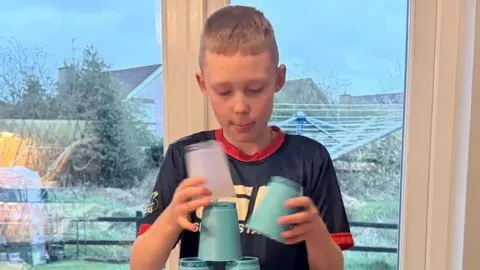Logan McCarroll is a boy with blonde hair, he is standing in front of a set of glass patio doors wearing a blue top and is stacking light blue cups in a pyramid. He is standing in a living room with white walls.