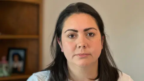 Rebecca Brown has shoulder-length, dark brown straight hair, tucked behind her ears, dark brown eyebrows and brown eyes. She is wearing a white top and looks resigned or disappointed. Behind her, and out of focus, is a white wall and a wooden bookshelf with a framed photograph on it.