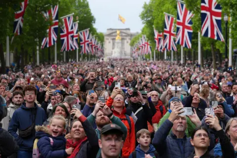 Reuters A huge crowd gather in the Mall after the procession, and look up to the sky while filming and taking photos with mobile phones, with union flags and green foliage lining the street and Buckingham Palace in the background.