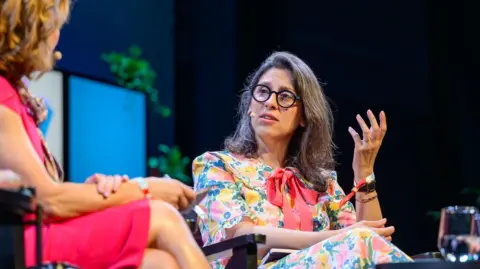 Sam Hardwick/Hay Festival A woman with dark hair and glasses wearing a bright multicoloured floral dress with a pink bow on the front. She's sat on a chair and is in conversation with another woman sat across from her, who is blonde and wearing a pink dress.