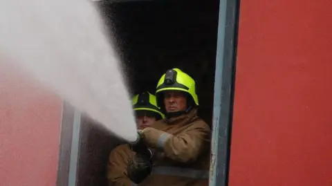Two firefighters in hi-vis helmets and fire jackets spraying a hose 