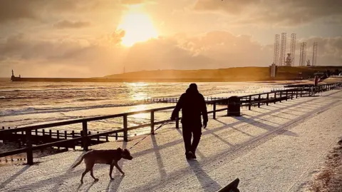 Sunrise over Aberdeen Beach. The sky is a pale yellow colour and the sun is reflecting on the water. A bench and a man walking a dog are in the foreground. There is frost on the ground.