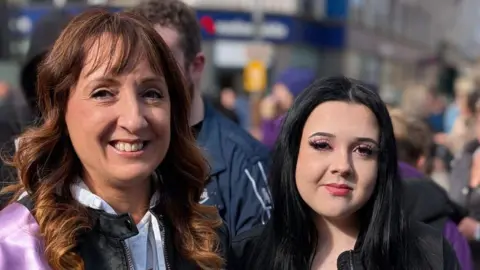 Lesley has long brown hair and is smiling. Emma has long black hair. They are standing next to each other.