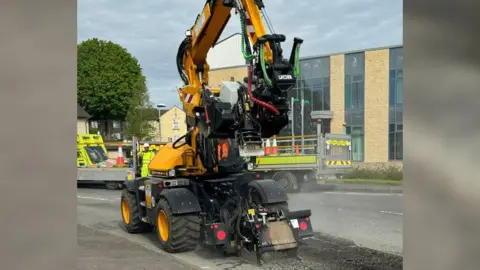 Gloucestershire County Council Yellow vehicle with tractor tyres and a pothole filling attachment on an arm