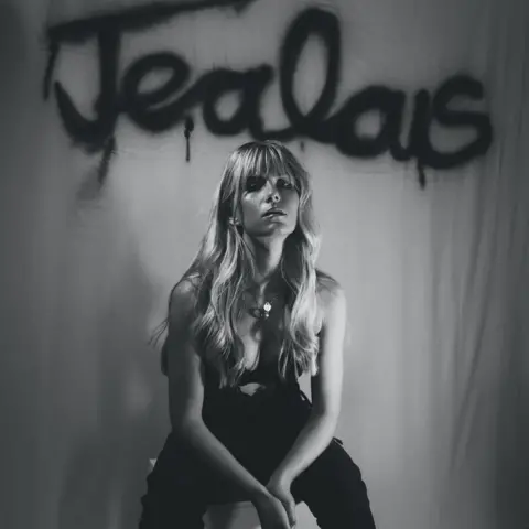 Romy Caton-Jones A black and white photo of a woman with long blonde hair, wearing black trousers and a top and sitting on a stool with her hands together between her legs. The word "jealous" is painted on a cloth draped down behind her.