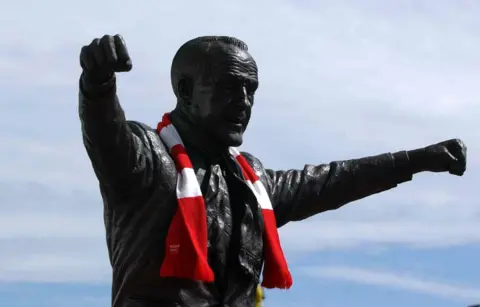 PA Media Close up shot of Bill Shankly's bronze statue outside Anfield shows the metal statue wearing a red and white Liverpool scarf, its arms stretched out as if to embrace the crowd. It sits against a cloud-streaked blue sky behind.