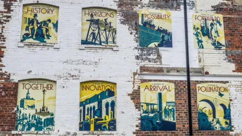 Stockton Council The mural is on the side of a brick building, and consists of eight individual panels that display words and images associated with the railway. Some of the words visible include: together, innovation, arrival, history, etc. Each panel is made up of shades of yellow and blue, with the text in red.