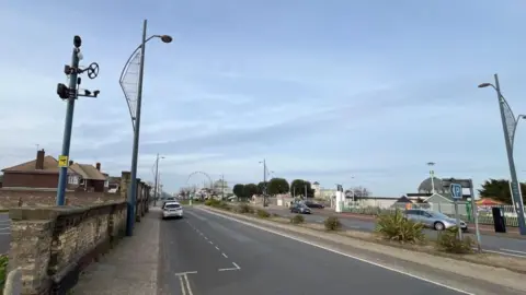 Andrew Turner/BBC Great Yarmouth's Marine Parade showing a two-lane road with plants separating each lane. Various sea front buildings and plants, with lamp standards each side, plus the camera post with the Accoustic Recognition Camera on the left-most of the picture.