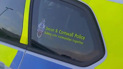 A close up image of a Devon & Cornwall Police car. The photo shows the rear back window of the car with the forces branding written on the window. The car has the usual police colours of yellow and blue printed on white. 