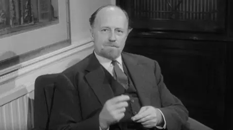 Black and white screengrab of a man in a three-piece suit sitting in an armchair and talking to the camera. He has balding, swept-back hair and is sitting in front of a wall with a radiator and picture to the left and a dark wooden cabinet behind him. 