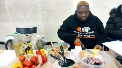 A woman in a black hoodie chopping vegetables with bowls of food and utensils in front of her