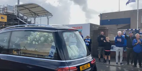 BBC A hearse - with Mr Keen's coffin and flowers inside - drives along a street. Fans are clapping and wearing blue and white striped scarves.