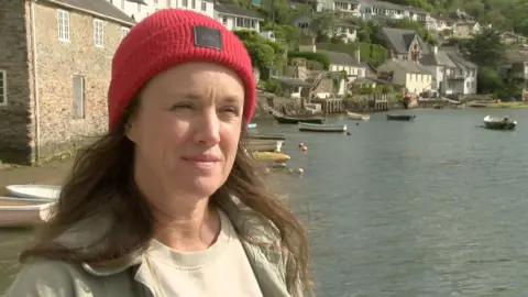 A woman stands by Newton Creek in South Devon. You can see small boats attached to buoys in the background. There are buildings along the edege of the river in the distance. 
