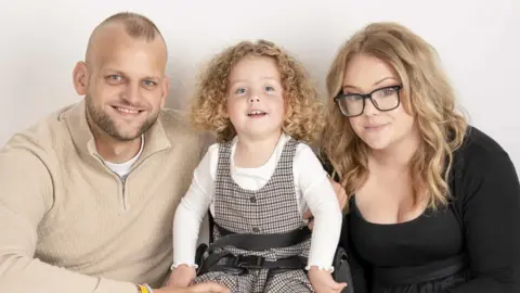 Forever Timeless Photography Tony and Carrie Pearson sitting outside with their daughter Grayce. Grayce is smiling widely at the camera and has shoulder-length, curly, blonde hair. She is wearing a white, long-sleeved top and a brown checked pinafore dress. Carrie is wearing a black top and skirt and has long, blonde hair and black-rimmed glasses. Tony has a shaved/bald head and a beard and is wearing a beige, long-sleeved top.
