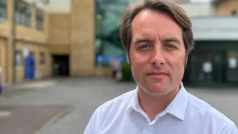 A head and shoulders picture of Thom Lafferty. He is standing in front of a hospital and is wearing a white shirt with the top button undone.