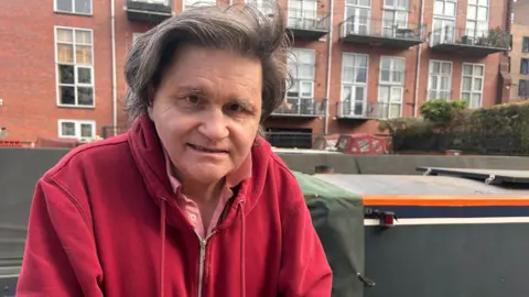 A man with wavy grey hair in a red coat with a large red brick building behind him