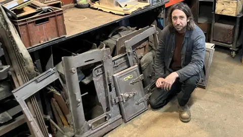 Olly Osborne crouches down alongside a dismantled old-fashioned oven. The metal pieces are laid against a workbench - which has other paraphernalia on it. He is wearing a grubby jacket and worn work shoes. 