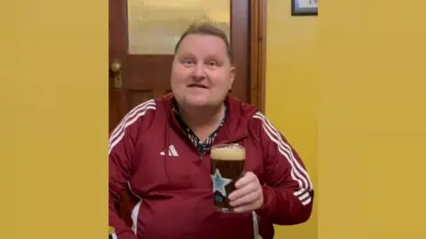 John Booth John Booth, mengenakan pakaian olahraga Adidas merah, dan memegang gelas pint. Dia melihat kamera dan memiliki rambut coklat muda pendek. Kaca memiliki logo Blue Star dari Newcastle Brown Ale.