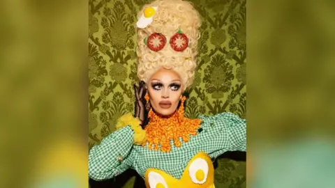 Knowsley Pride Sister Sister wears a green gingham dress with yellow eggs on the front and a large orange collar. Their hair is styled very high and is blond and curled with egg and tomato embellishments