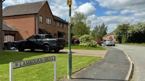 BBC A curved road with a sign in the foreground reading Lambourn Drive and a number of houses stood behind lawns on the left hand side