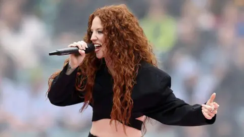 Getty Images Jess Glynne is singing into a mic, she is wearing a cropped black suit jacket. SHe has long curly ginger hair. 