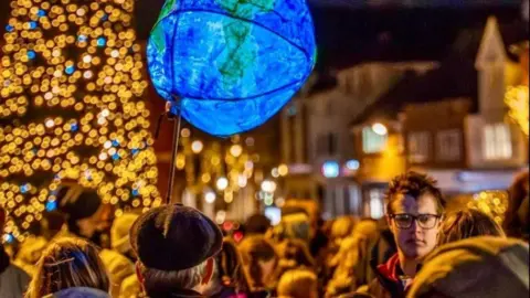 Banbury BID Blurred heads in a crowd with a lantern in the shape of the Earth held aloft and the gold and blue lights of a Christmas tree to left.