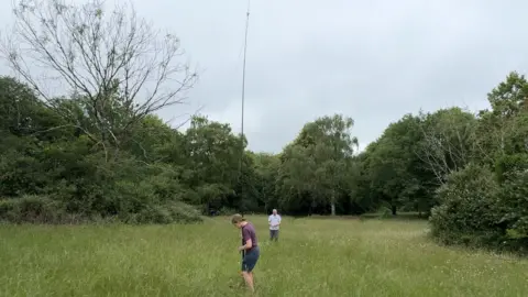 A woman and a man in a field assembling a radio contact point. The are using a large tall black pole and dangling long antenna wires from the top of it to the ground.