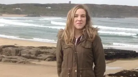 PA Media A woman with blonde hair and wearing a brown coat is standing on a beach in front of sand and rocks. The sea is in the background.
