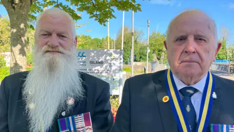 Paddy, left, is looking at the camera and has a very long grey beard. He is wearing military uniform and has a number of medals on his left lapel. Brian, right, is looking at the camera. He is bald and also wearing military dress. He has a blue and yellow ribbon around his neck and also has a number of medals on his left lapel.