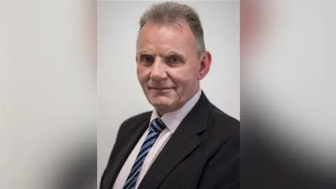 Somerset Council Alistair Hendry is wearing a black suit and blue striped tie. He is smiling at the camera. He is standing in front of a grey background.