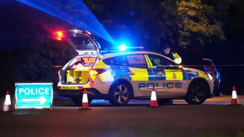 A police car parked at night with its blue lights on and an officer in a high-vis jacked stood at the front. It's boot is open and an LED sign reads 'closed'. There are cones and a blue police sign blocking the road.