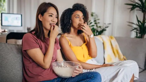 Getty Images Stock photo shows two women sitting on the sofa eating popcorn from a bowl with a desktop computer and plants in the background.
