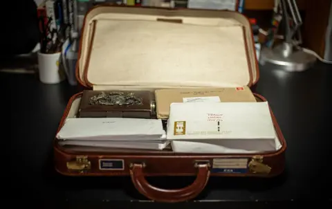 Charlie Northcott/BBC An open brown leather suitcase sits on a dark surface, containing a decorative metal box, a brown cardboard box, and several white envelopes. One envelope is marked 'FRAGILE HANDLE WITH CARE' in red text, and another has an address label. In the background, there are books, a cup holding pens and scissors, and a desk lamp.