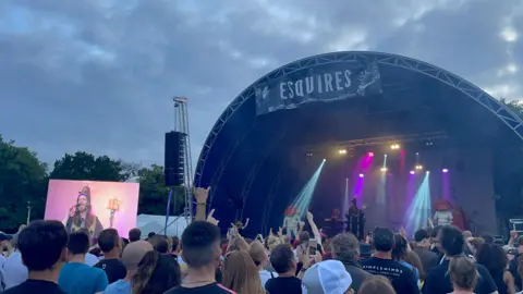 Bedford Borough Council A large group of people in front of a stage, with an act performing on it. A screen is to the left. Some people have put their arms up in the air. The sky is turning dark in the sky.
