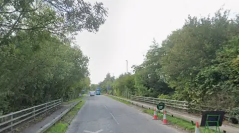 A large road with railings and trees on either side. Some traffic cones can be seen to the right of the carriageway.