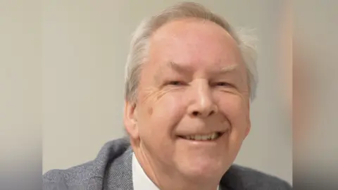 Jamie Niblock/BBC Chris Cobb smiles at the camera. He has short, thinning, grey hair. He wears a grey suit with a white shirt underneath. 